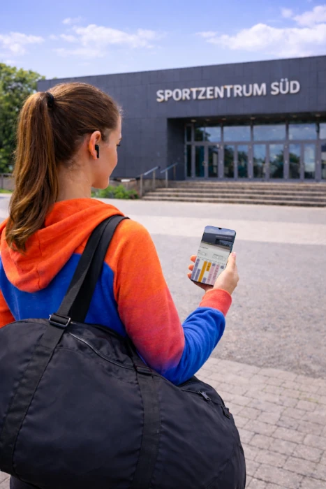 Eine Frau von hinten mit einer Sporttasche steht vor einem Sportzentrum und hat auf ihrem Smartphone das Sportstättenportal von Groundkeeper geöffnet